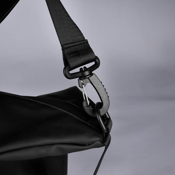Detailansicht einer schwarzen Reisetasche aus glattem Material. Im Fokus steht der Karabiner des Schultergurtes, der in eine Öse an der Seite der Tasche gehakt ist.