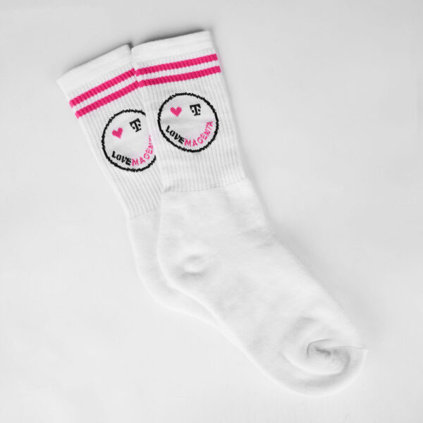 Image of the SMILEY SOCKS, both socks are offset on top of each other, you can see the two magenta stripes below the cuff and the smiley, whose right eye is a magenta heart and whose left eye is a black T logo. The mouth is formed by the lettering “LOVE MAGENTA”, with the word ‘LOVE’ woven in black and the word “MAGENTA” in magenta.