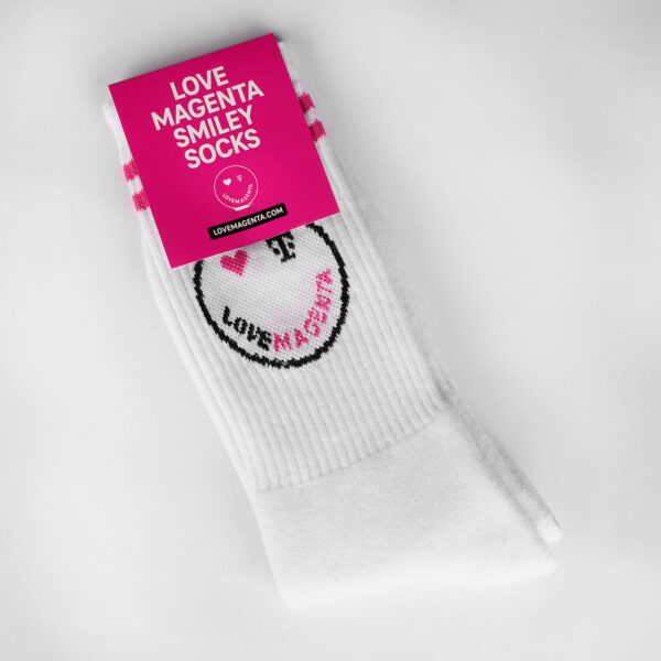 Image of the SMILEY SOCKS with a magenta-colored band that reads “LOVE MAGENTA SMILEY SOCKS” and the size in white lettering. You can see the two magenta stripes below the cuff and the smiley, whose right eye is a magenta heart and whose left eye is a black T logo. The mouth is formed by the lettering “LOVE MAGENTA”, with the word ‘LOVE’ woven in black and the word “MAGENTA” in magenta.