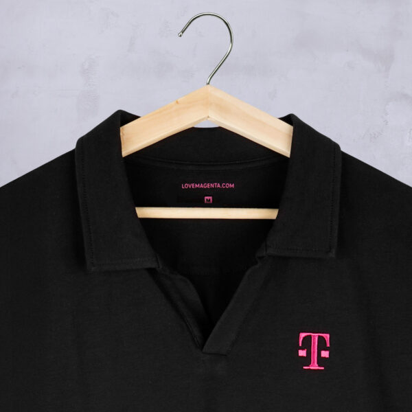 Detailed view of the BLACK POLO with focus on the collar. The polo shirt hangs on a wooden hanger. The buttonless seamed V-neck and the magenta-colored embroidered T-logo are visible