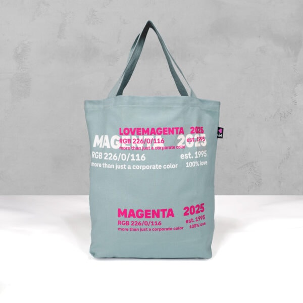 Eine hellblau-graue MAGENTA 2025 SHOPPER Tragetasche mit Henkeln steht aufrecht vor einem passenden Hintergrund. Sie trägt den fetten magentafarbenen und weißen Schriftzug "MAGENTA 2025" und den Satz "mehr als nur eine Unternehmensfarbe".