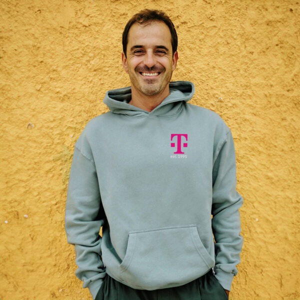 Ein lächelnder Mann, der den MAGENTA DEFINITION HOODIE mit magentafarbenem Schriftzug trägt, steht vor einer strukturierten gelben Wand, die Hände in den Taschen.