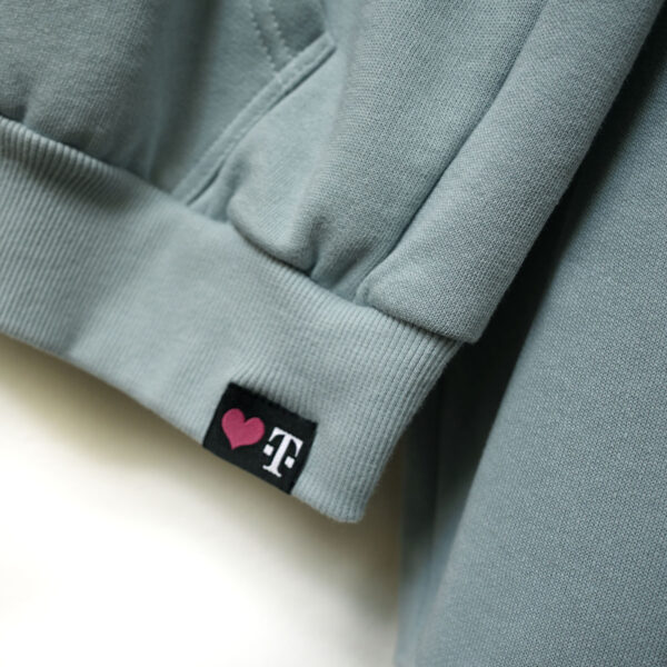 Eine Nahaufnahme eines graublauen MAGENTA DEFINITION HOODIE-Saums mit einem schwarzen Etikett, das ein magentafarbenes Herz neben einem weißen T zeigt, ein subtiles, stilvolles Detail.