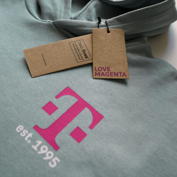 Der graublaue MAGENTA DEFINITION HOODIE mit einem großen magentafarbenen T-Logo mit "est. 1995" aufgedruckt, begleitet von zwei braunen Anhängern, einer mit "LOVE MAGENTA" in magentafarbener Schrift.