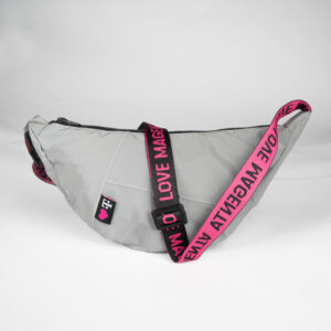 The UPCYCLED SHOULDER BAG REFLECTIVE is a gray reflective bag with an adjustable strap in magenta and black with the lettering “LOVE MAGENTA”, a reflective surface, a zipper and a heart patch and the T logo on the front.