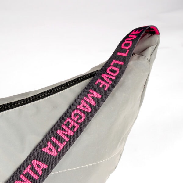 Eine Nahaufnahme der UPCYCLED SHOULDER BAG REFLECTIVE in Grau mit reflektierendem schwarzem Reißverschluss und schwarzem Riemen mit magentafarbenem "LOVE MAGENTA"-Schriftzug auf weißem Hintergrund.