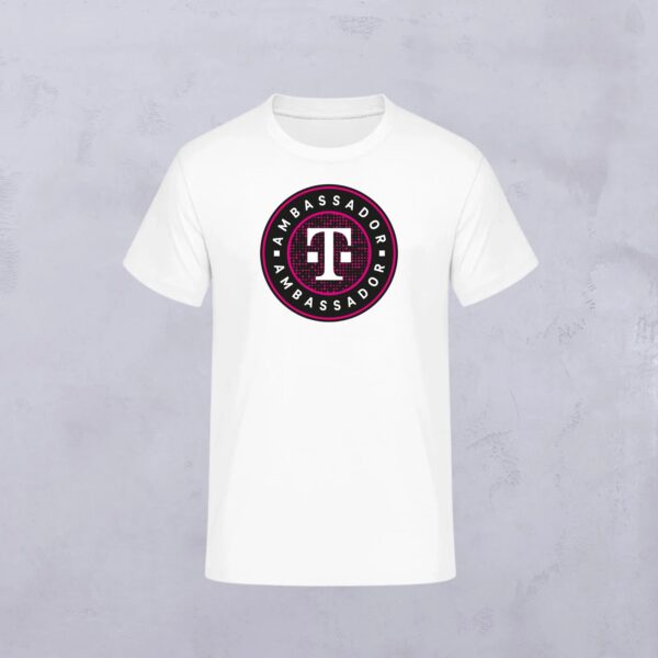 Das AMBASSADOR ROUND T-SHIRT WHITE zeigt ein kreisförmiges "AMBASSADOR"-Logo mit dem T-Logo in der Mitte auf einem weißen T-Shirt vor einem hellgrauen Hintergrund.