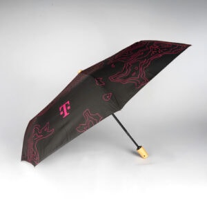 Der schwarze Regenschirm T WEATHER GUARD mit Holzgriff, magentafarbenem abstraktem Linienmuster und magentafarbenem Logo auf dem Schirmdach, dargestellt vor einem schlichten weißen Hintergrund.