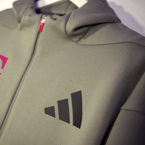 A close-up of the ADIDAS T ZIP JACKET UNISEX in the color clay with hood and black Adidas logo on the left chest and magenta T logo on the right chest.