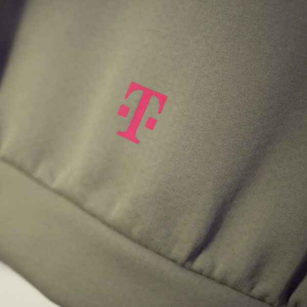 A close-up of the fabric in the clay color of the ADIDAS T ZIP JACKET UNISEX shows a small, centered T logo in magenta.