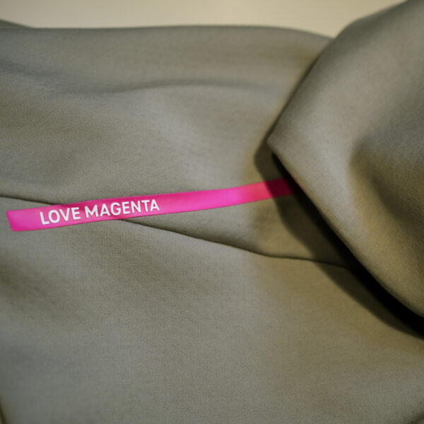 The clay-colored fabric of the ADIDAS T ZIP JACKET UNISEX is gently pleated and features a magenta stripe across the shoulder seam with “LOVE MAGENTA” printed in white letters.