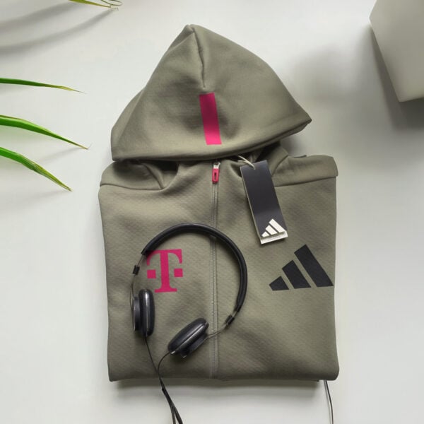 The folded ADIDAS T ZIP JACKET UNISEX with a magenta-colored label, the text “LOVE MAGENTA” and a black Adidas logo lies on a white surface. Black headphones and cardboard tags are attached to the jacket.