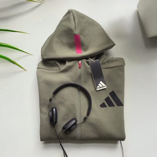 Eine gefaltete olivgrüne ADIDAS ZIP JACKET UNISEX mit schwarzem Logo, magentafarbenem Etikett und hochgezogener Kapuze liegt auf einer weißen Fläche neben schwarzen Kopfhörern, in deren Nähe grüne Pflanzenblätter und ein weißer Übertopf teilweise sichtbar sind.