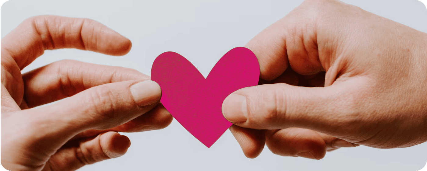 Zwei Hände halten sanft ein kleines, magentafarbenes Papierherz vor einem hellen, neutralen Hintergrund zusammen.