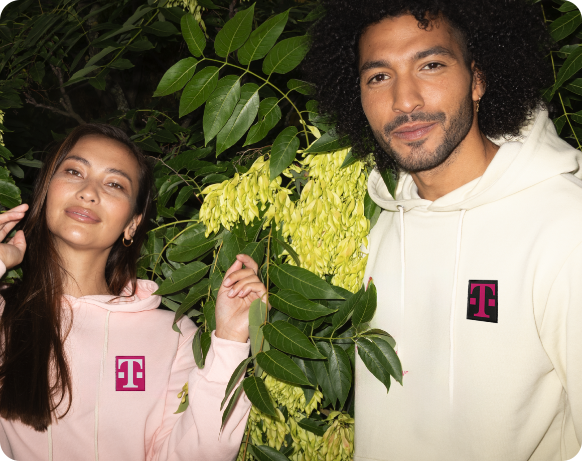 Eine Frau und ein Mann in hellen Kapuzenpullis mit Magenta T-Logos, lächelnd nebeneinander vor üppigen Grünpflanzen. LOVE MAGENTA Partner & Projekte.