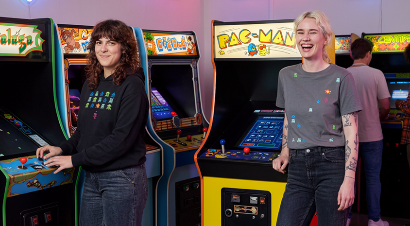 Two people are enjoying classic arcade games, including Pac-Man; one is wearing a LOVE MAGENTA T-shirt. Two others are hugging behind them, celebrating GAME ON moments.