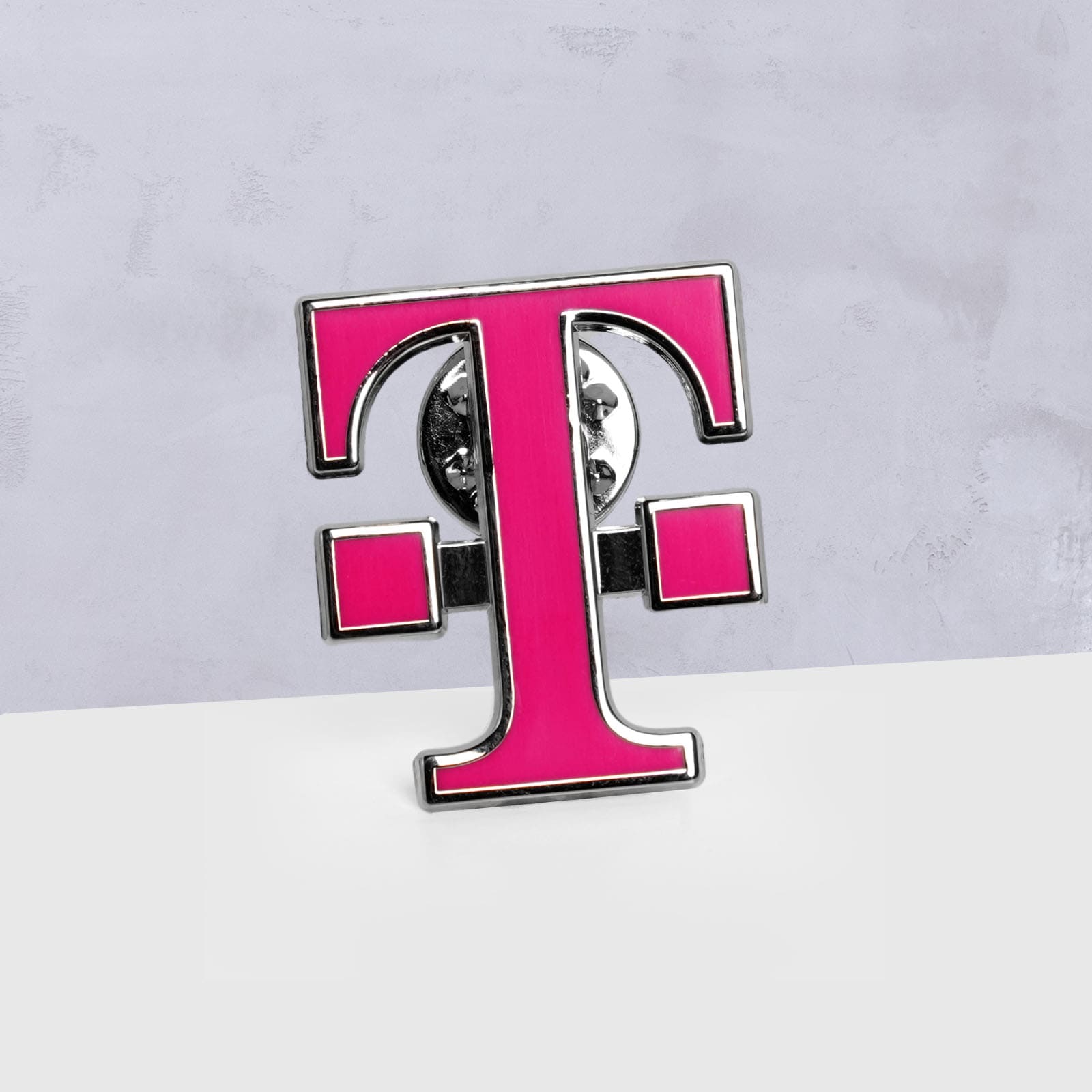 Eine rosa-silberne Anstecknadel in Form des Buchstabens T mit zwei kleinen Quadraten auf jeder Seite, vor einem grau-weißen Hintergrund.