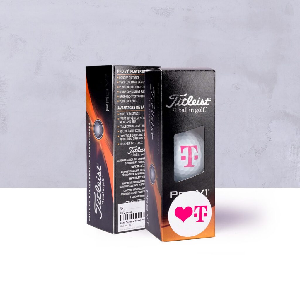 Zwei Schachteln mit Titleist Pro V1-Golfbällen, von denen eine geöffnet ist und einen Ball mit einem rosa T-Logo und einem Herzaufkleber auf der Schachtel zeigt. Der Hintergrund ist hellgrau, und die Schachteln stehen aufrecht.