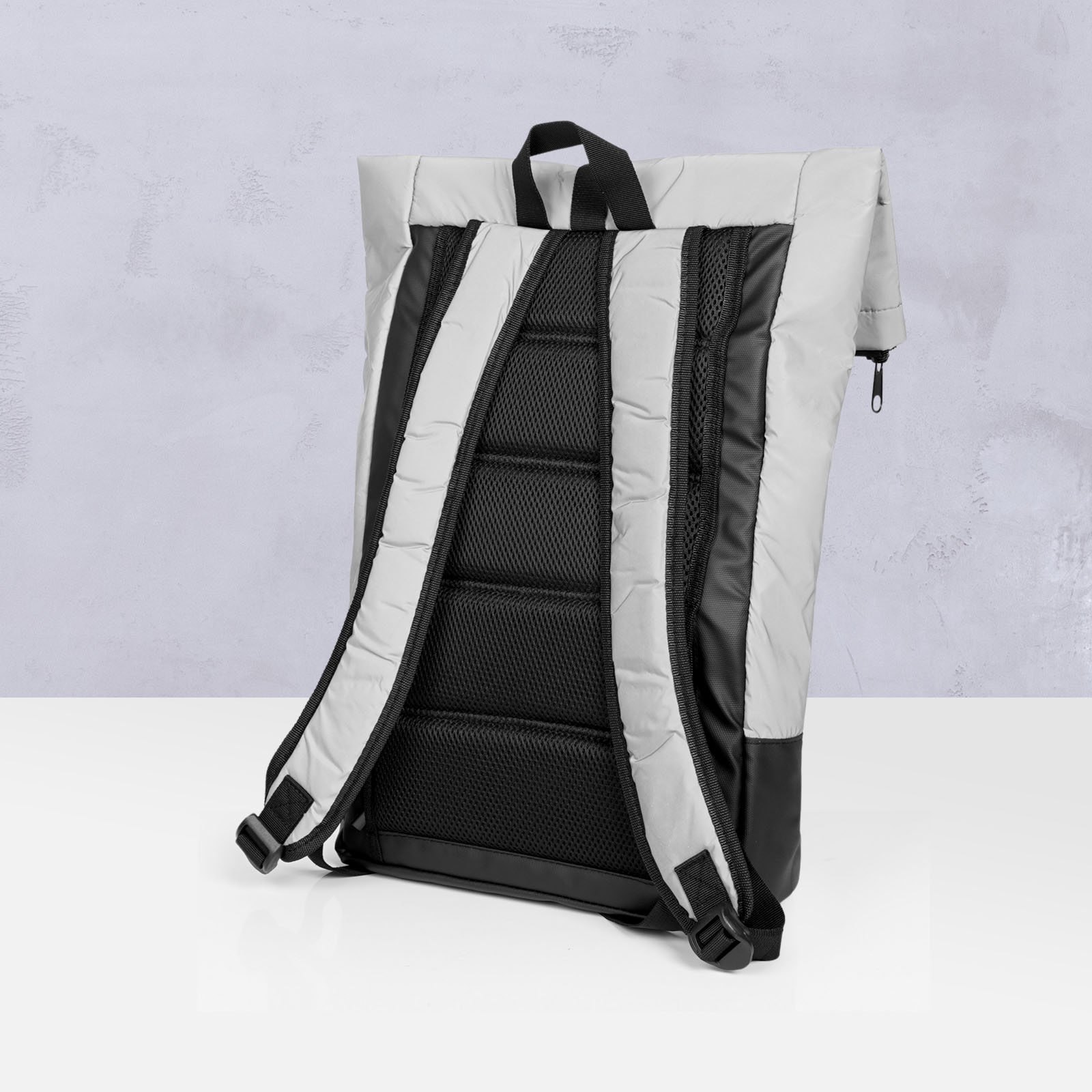 Ein moderner, grau-schwarzer Rucksack ist aufrecht auf einer weißen Fläche abgebildet; vor einem hellen, strukturierten Hintergrund sind die gepolsterten Schultergurte und ein Tragegriff an der Oberseite zu sehen.