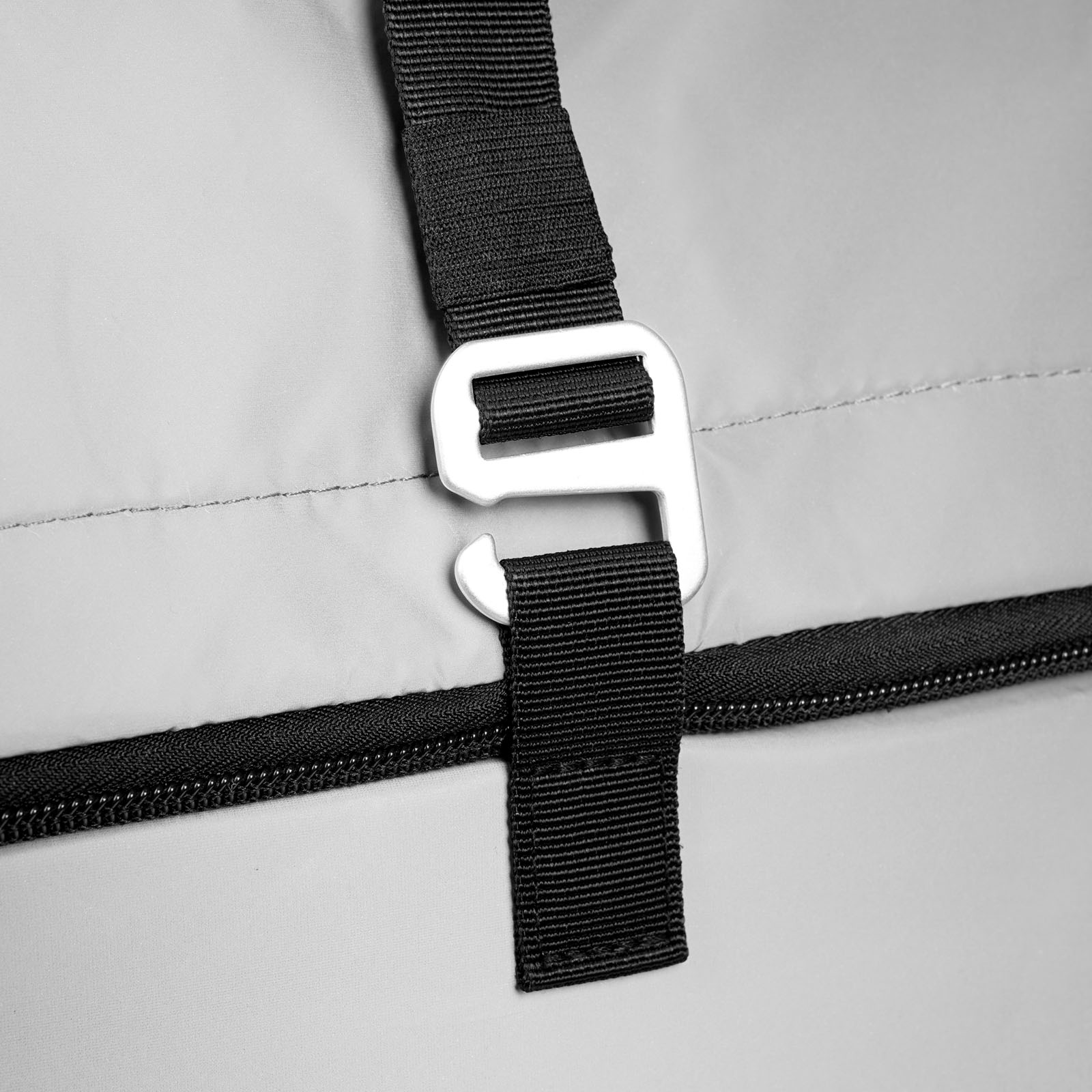 Nahaufnahme eines schwarzen Riemens mit einem silbernen Metallhaken, der an einer hellgrauen Stofftasche befestigt ist. Unterhalb des Riemens verläuft horizontal ein schwarzer Reißverschluss, und auf dem Stoff sind Nähte zu erkennen.