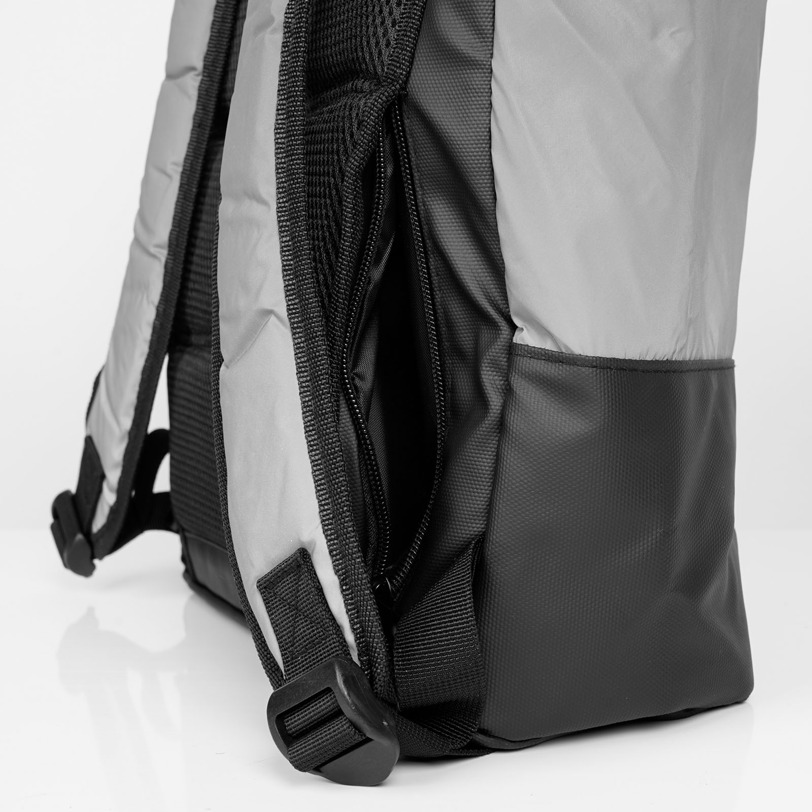 Nahaufnahme einer Seitenansicht eines grau-schwarzen Rucksacks, die eine Seitentasche mit Reißverschluss, einen gepolsterten Schultergurt und eine Kunststoffschnalle vor weißem Hintergrund zeigt.