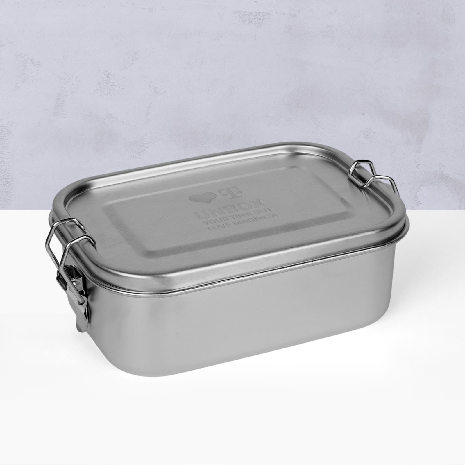 Eine rechteckige Lunchbox aus Edelstahl mit einem fest schließenden Deckel und Metallverschlüssen an beiden Seiten, platziert auf einer weißen Fläche mit hellgrauem, strukturiertem Hintergrund.