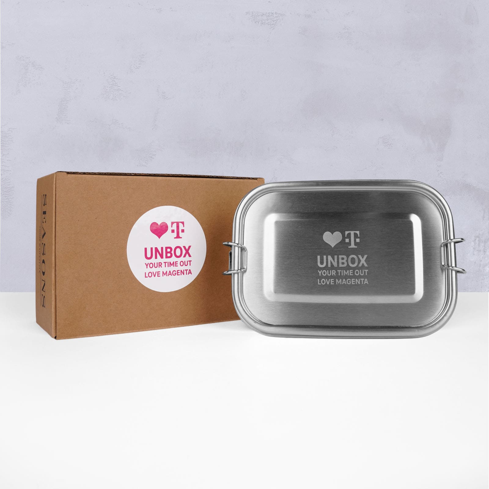Eine Metall-Brotdose mit der Aufschrift „UNBOX YOUR TIME OUT LOVE MAGENTA“ und einem Logo befindet sich neben einem braunen Karton mit einem passenden Aufkleber vor einem hellgrauen Hintergrund.