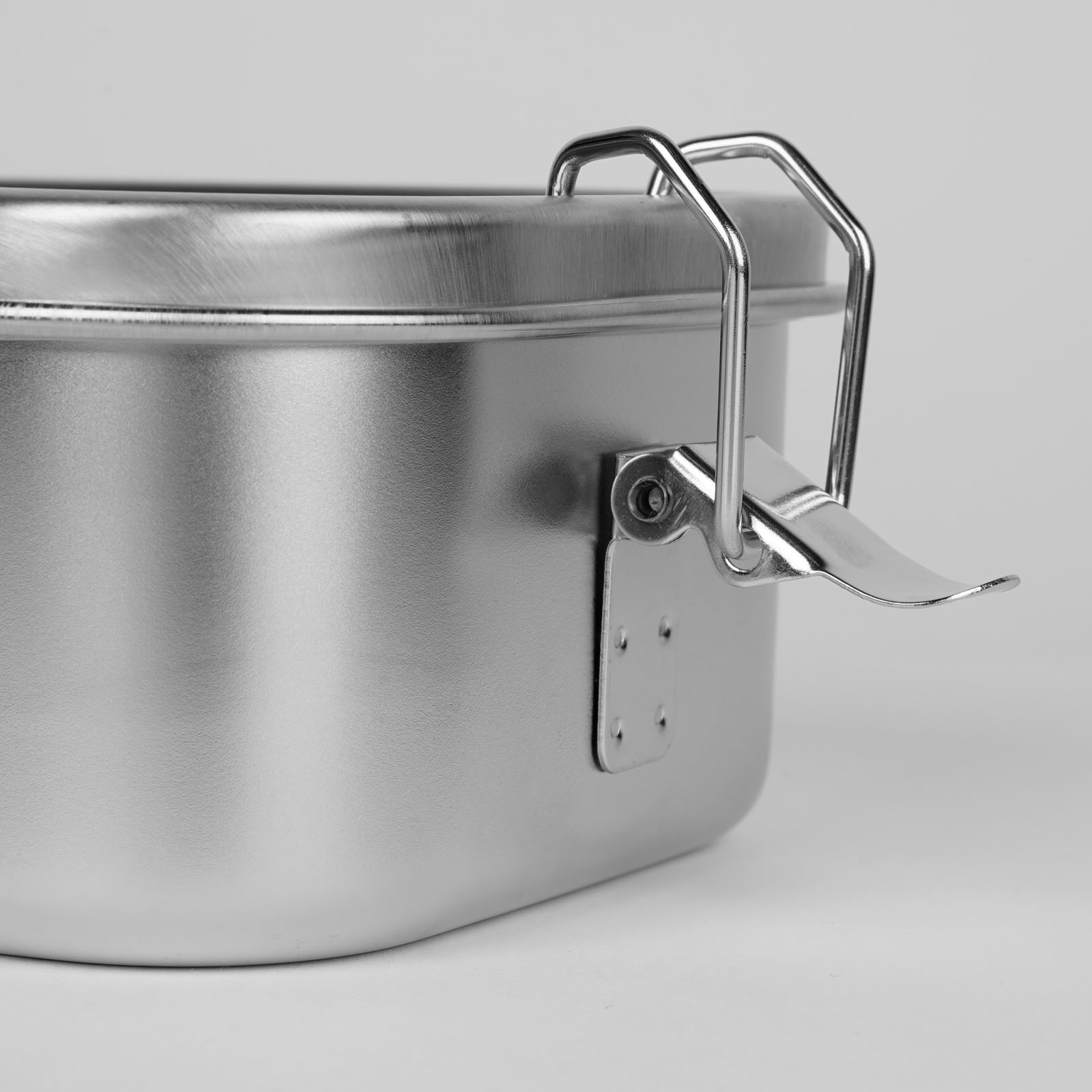 Eine Nahaufnahme eines Metallbehälters mit Deckel, dessen Verschluss durch einen hakenförmigen Metallverschluss gesichert ist. Der Behälter hat eine glatte, matte Oberfläche und wurde vor einem einfarbigen, hellen Hintergrund fotografiert.