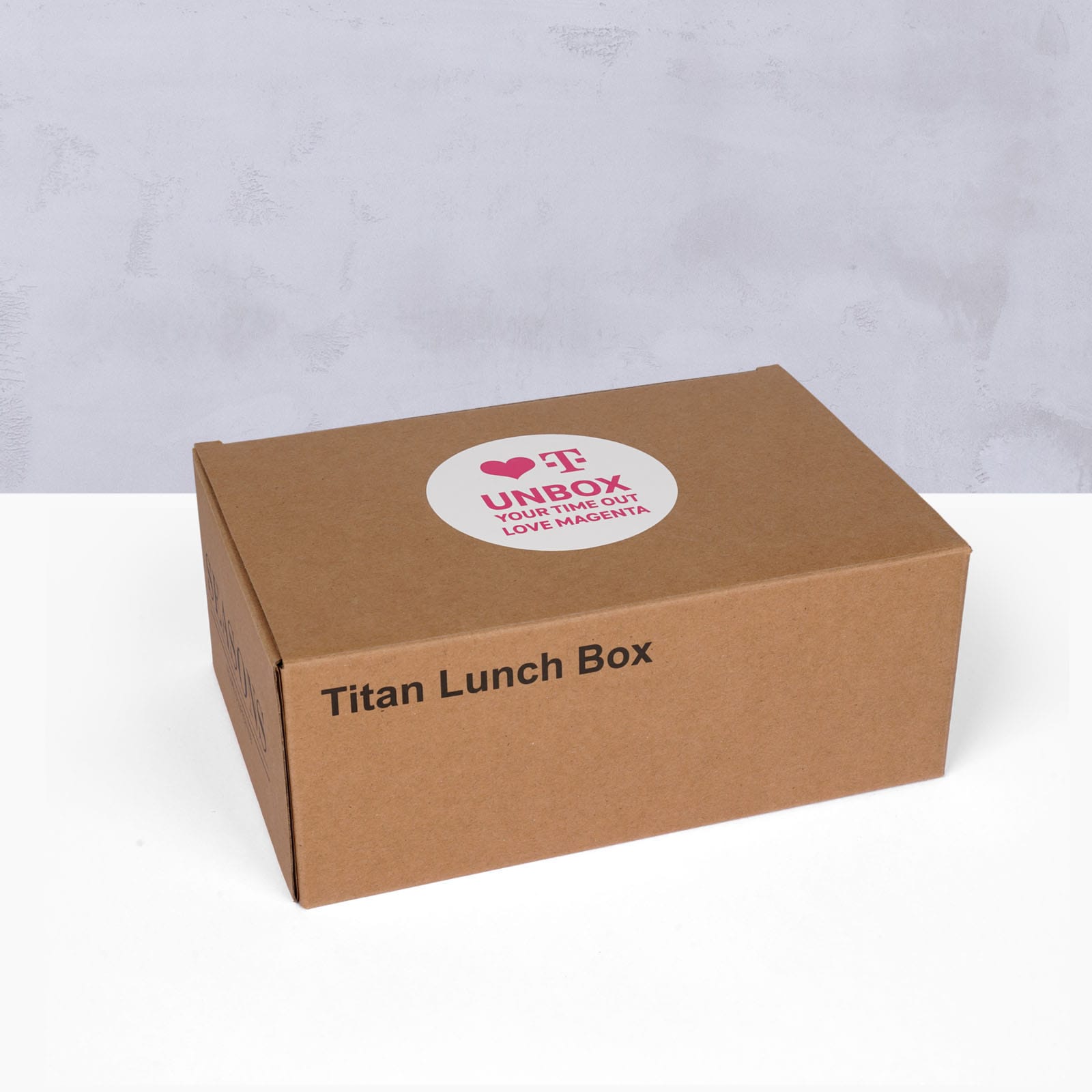 Auf einer weißen Fläche vor hellgrauem Hintergrund steht ein brauner Karton mit der Aufschrift „Titan Lunch Box“ und einem runden Aufkleber oben, auf dem zu lesen ist: „UNBOX YOUR TIME OUT. LOVE HAPPENS.“.