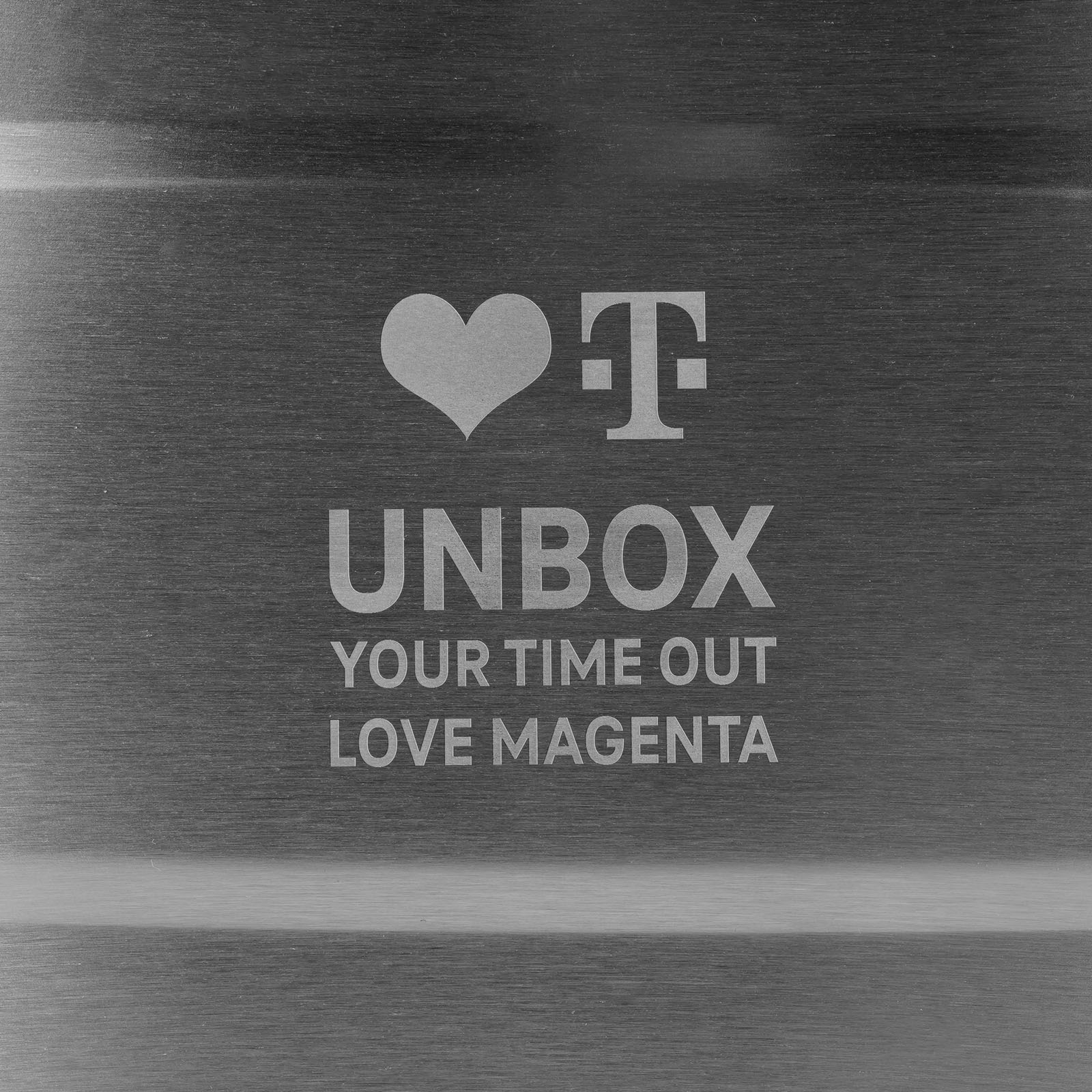 Oberfläche aus gebürstetem Metall mit einem Herz, dem T-Mobile-Logo und dem Schriftzug: UNBOX YOUR TIME OUT LOVE MAGENTA.