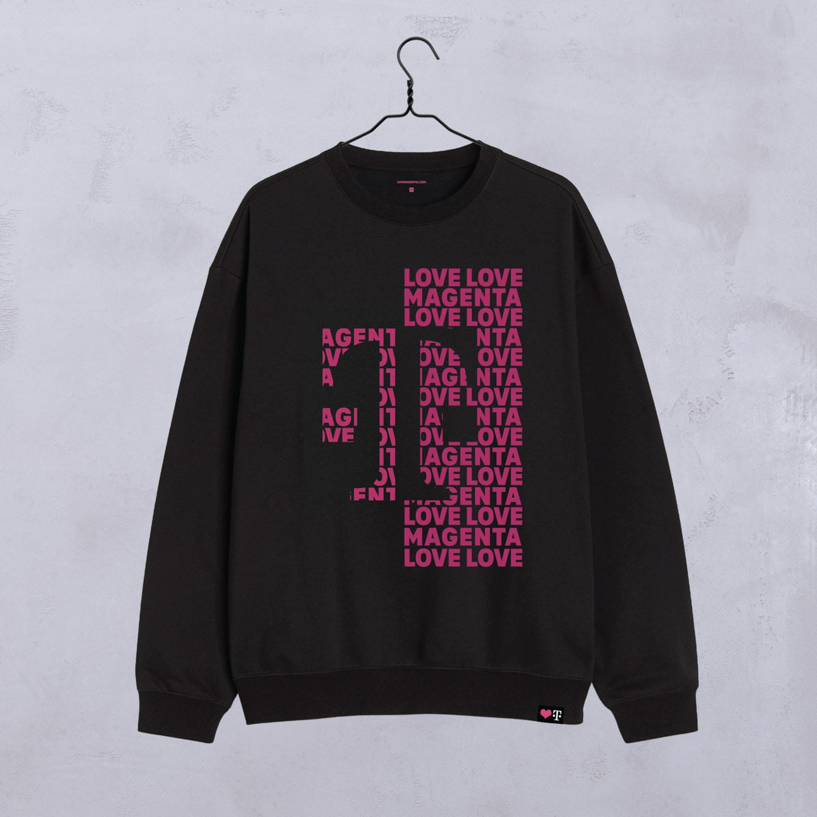 Auf einem Kleiderbügel hängt ein schwarzes Sweatshirt mit einer auffälligen rosa Aufschrift in Form eines großen M, wobei die Worte „LOVE LOVE MAGENTA“ auf der Vorderseite wiederholt sind. Der Hintergrund ist eine schlichte, hellgraue Wand.