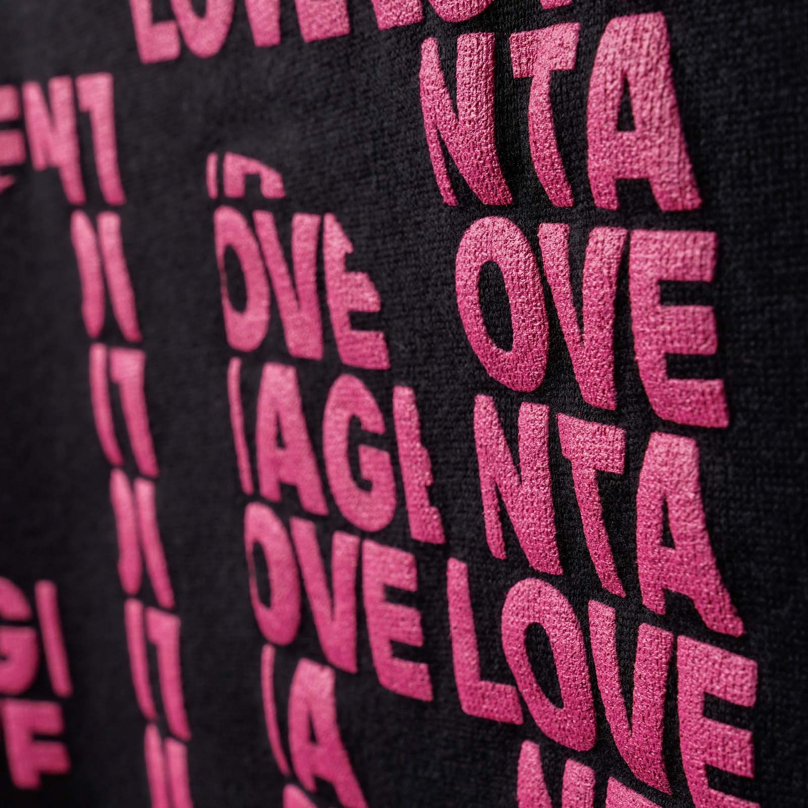 Nahaufnahme von rosa, fetter, erhabener Schrift, auf der wiederholt „LOVE MAGENTA“ auf schwarzem Stoff gedruckt ist, wobei einige Buchstaben teilweise abgeschnitten sind oder sich überlappen.