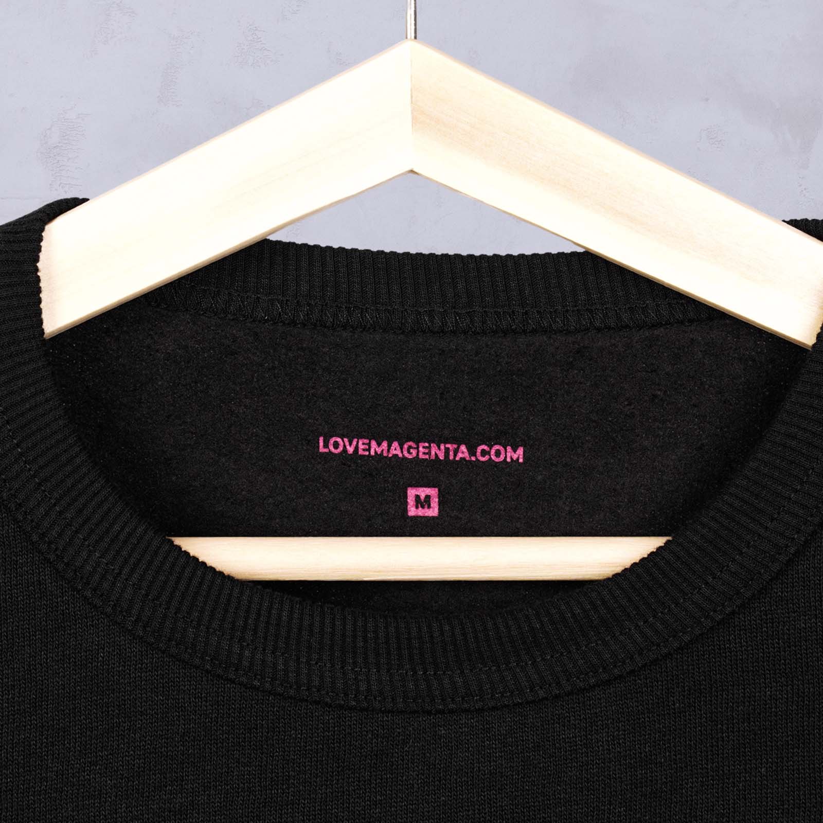 Eine Nahaufnahme eines schwarzen Pullovers auf einem Holzkleiderbügel, auf der im Kragenbereich ein Etikett zu sehen ist, auf dem in rosa Schrift „LOVEMAGENTA.COM“ und ein „M“ stehen.