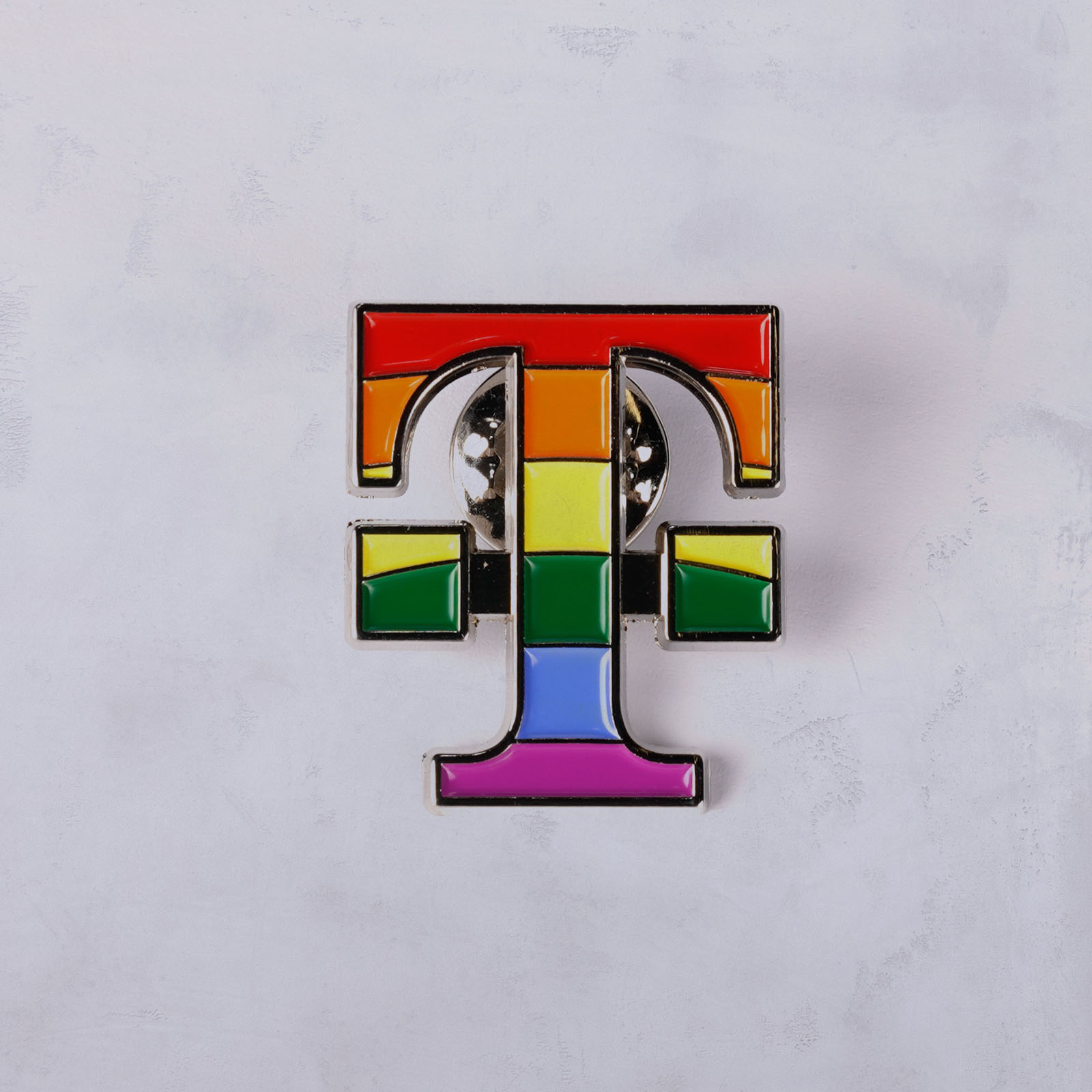 Ein Anstecker in Form des Buchstabens T, verziert mit horizontalen Regenbogenstreifen, die den LGBTQ+-Stolz symbolisieren, auf einem hell melierten Hintergrund.