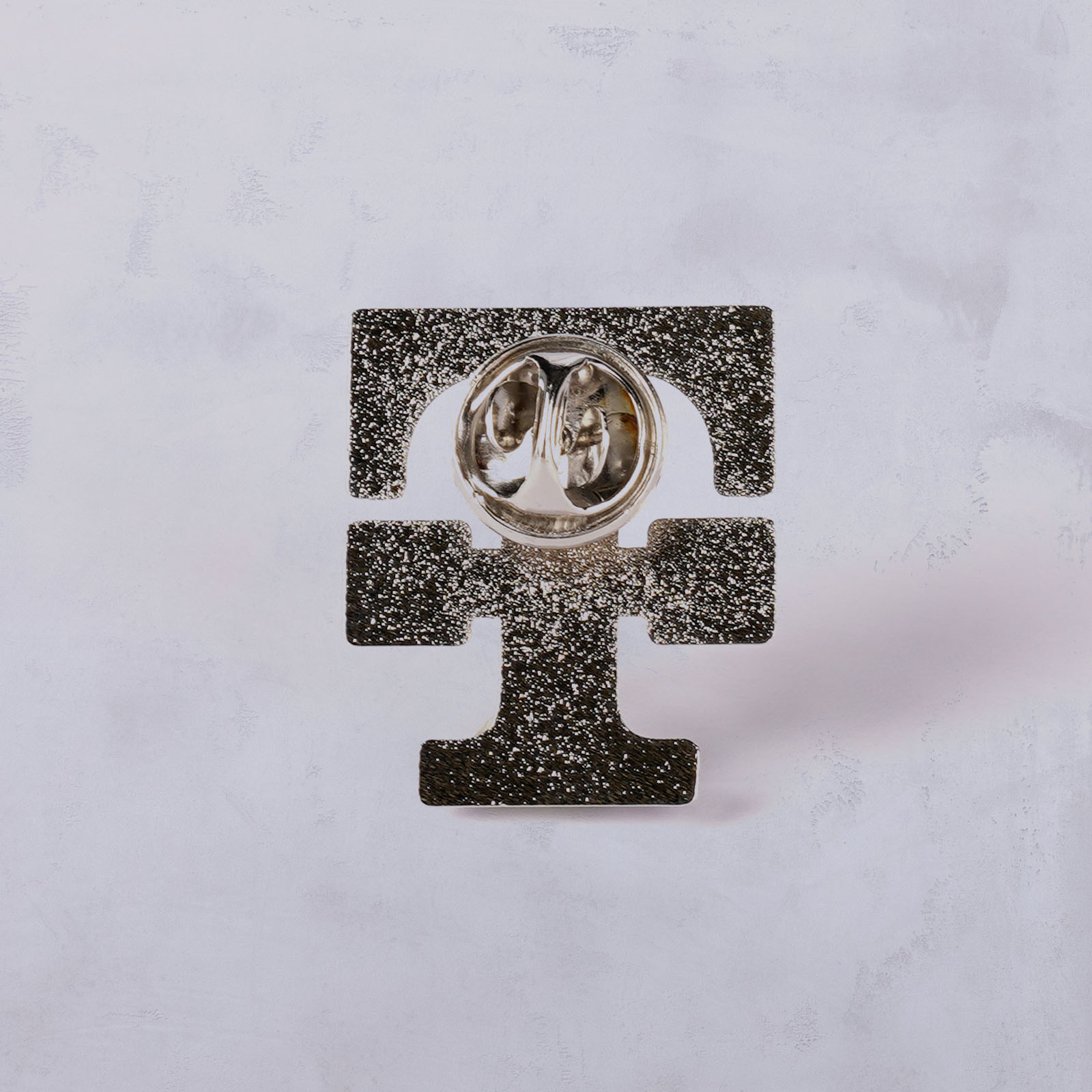 Ein metallischer Gegenstand in Form eines markanten, stilisierten Buchstabens E mit einem runden, reflektierenden Element in der Mitte, vor einem hellen, strukturierten Hintergrund.