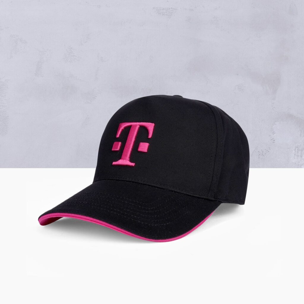 Eine schwarze Baseballkappe mit einem leuchtend pinkfarbenen „T“-Logo auf der Vorderseite und einem pinkfarbenen Rand an der Krempe, vor einem hellgrauen Hintergrund.