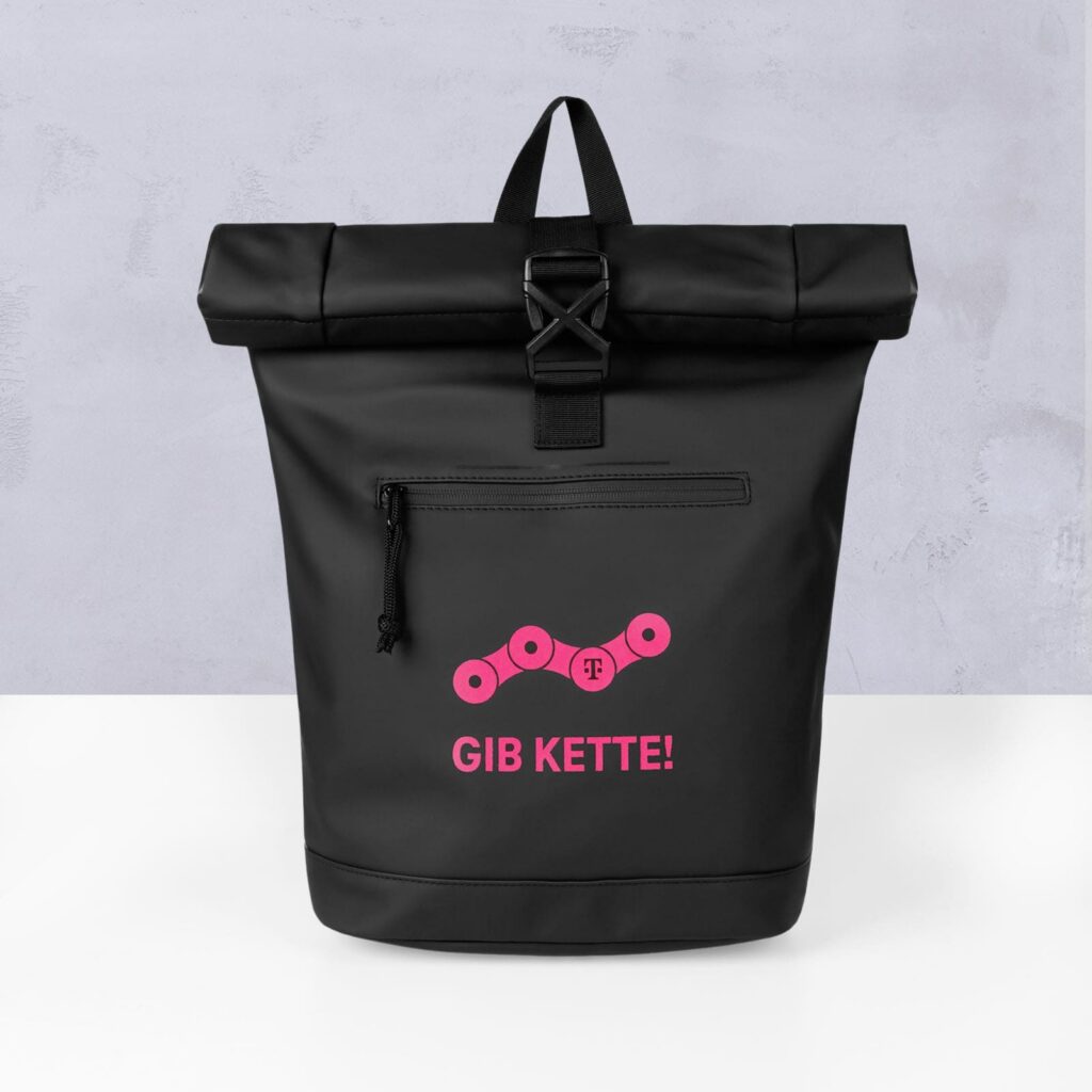 Ein schwarzer Rucksack mit Rollverschluss und einer Reißverschlusstasche vorne, auf dessen Vorderseite ein rosa Fahrradkettenmotiv und der Schriftzug „GIB KETTE!“ zu sehen sind, vor einer hellvioletten, strukturierten Wand.