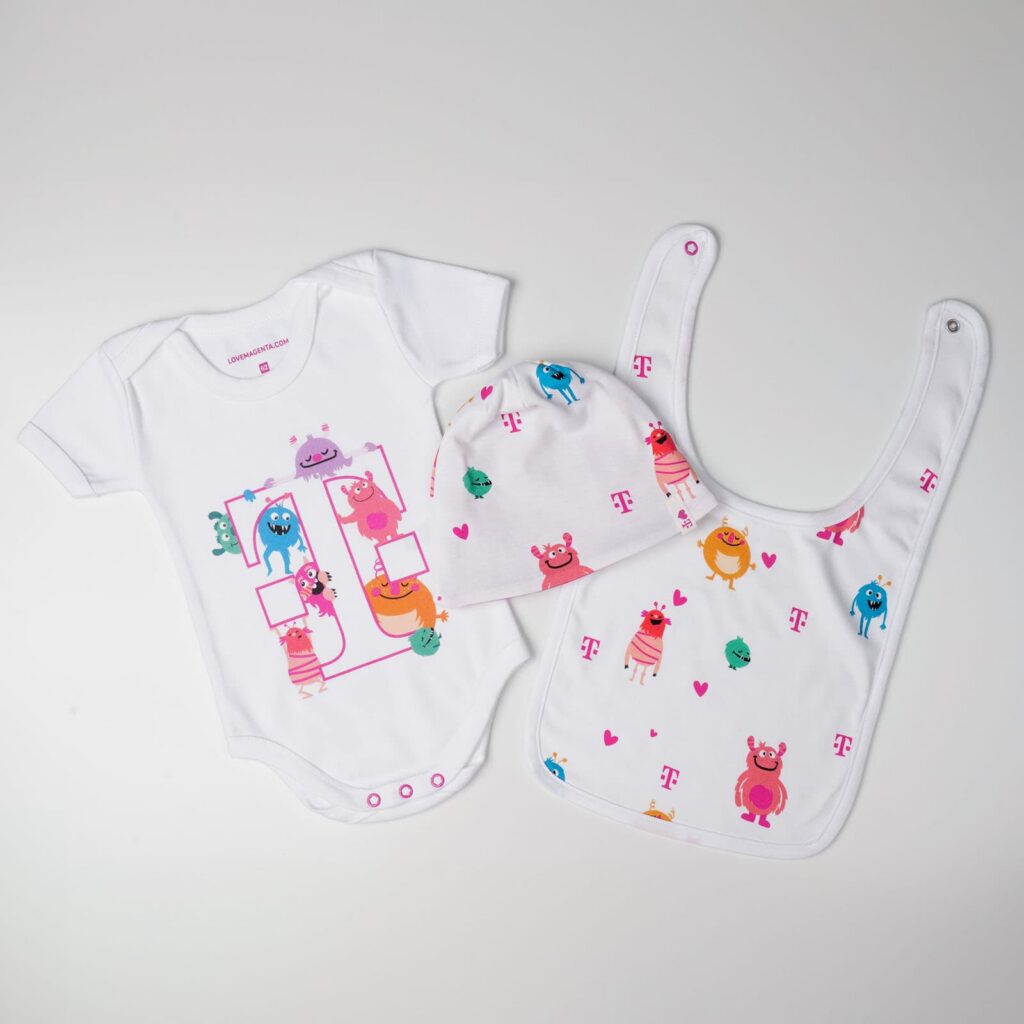 Set aus weißem Baby-Strampler, Mütze und Lätzchen mit bunten Comic-Monstern und rosa T-Mobile-Logos auf weißem Hintergrund.
