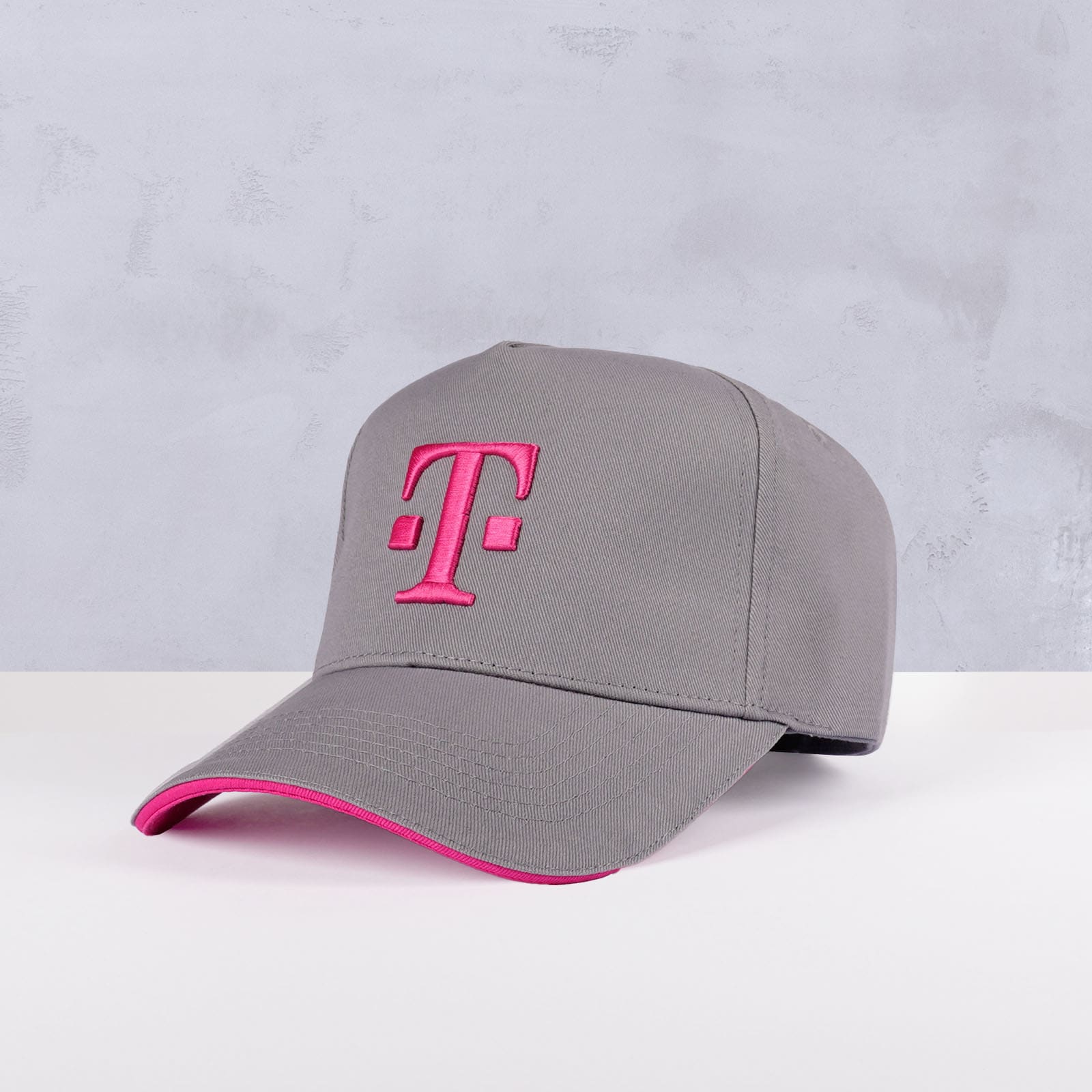 Eine graue Baseballkappe mit einem rosa gestickten T-Logo auf der Vorderseite und einem rosa Rand entlang der Krempe, vor einem hellgrauen Hintergrund.