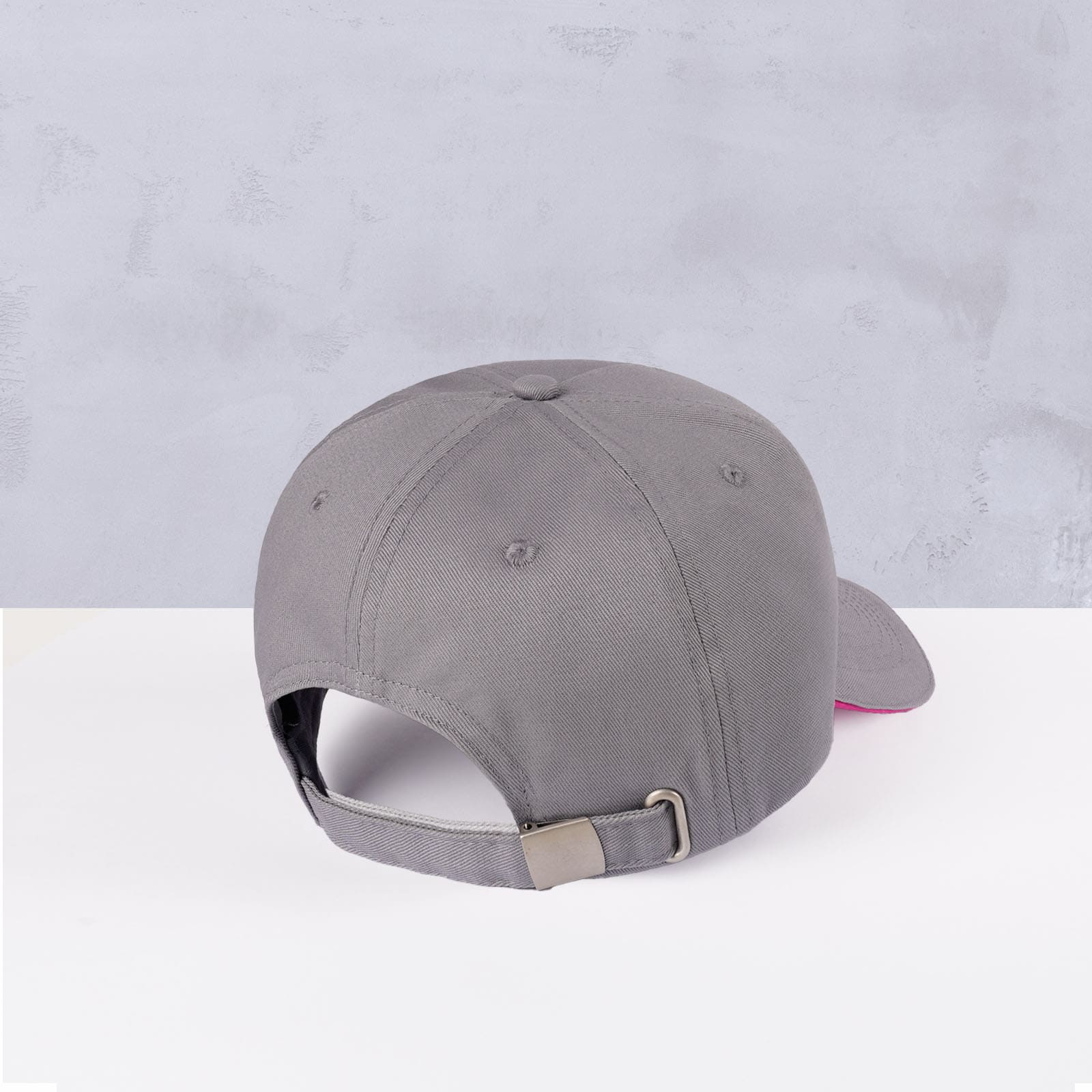 Eine graue Baseballkappe, von hinten gezeigt, mit einer Metallschnalle und einem verstellbaren Riemen, auf einer hellen Unterlage mit hellgrauem, strukturiertem Hintergrund platziert.