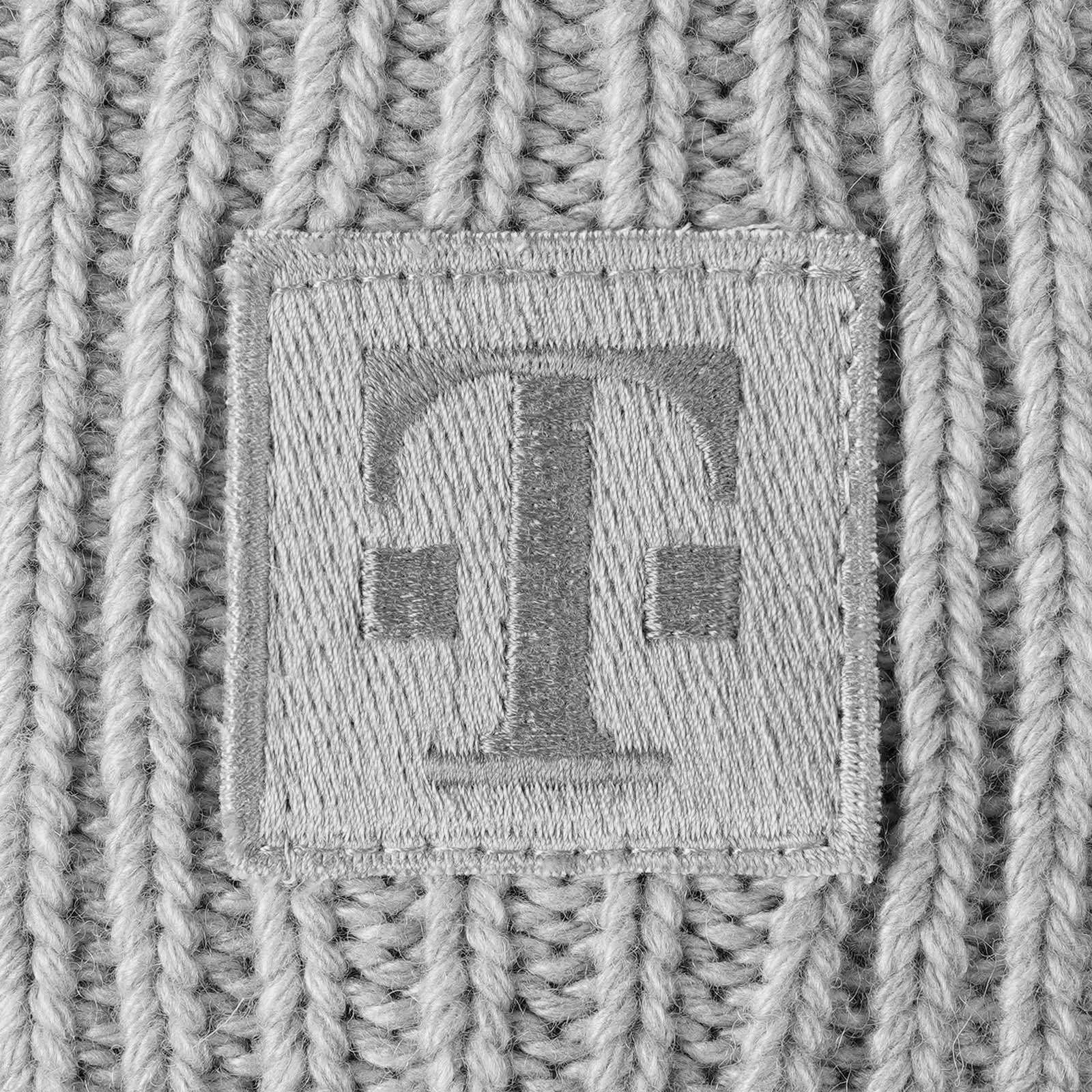 Eine Nahaufnahme eines grauen Strickstoffs mit einem quadratischen Aufnäher, in dessen Mitte ein gesticktes „T“-Symbol zu sehen ist. Die Stickerei und der Stoff wirken strukturiert.