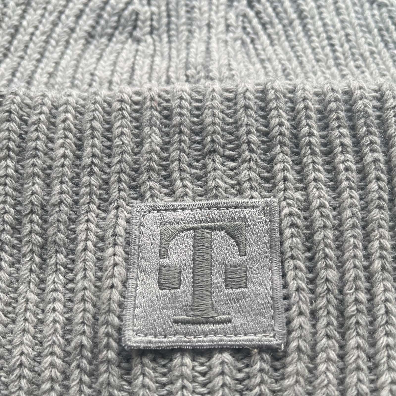 Eine Nahaufnahme eines grauen Strickstoffs mit einem quadratischen Aufnäher, auf dem ein gestickter Buchstabe „T“ aus farblich passendem grauen Garn zu sehen ist.