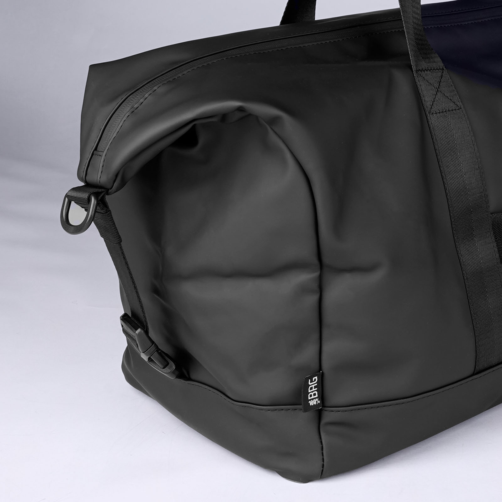 Eine Nahaufnahme einer schwarzen Reisetasche mit schwarzen Trägern und einem kleinen „BRC“-Etikett auf der Vorderseite, vor einem schlichten hellen Hintergrund.