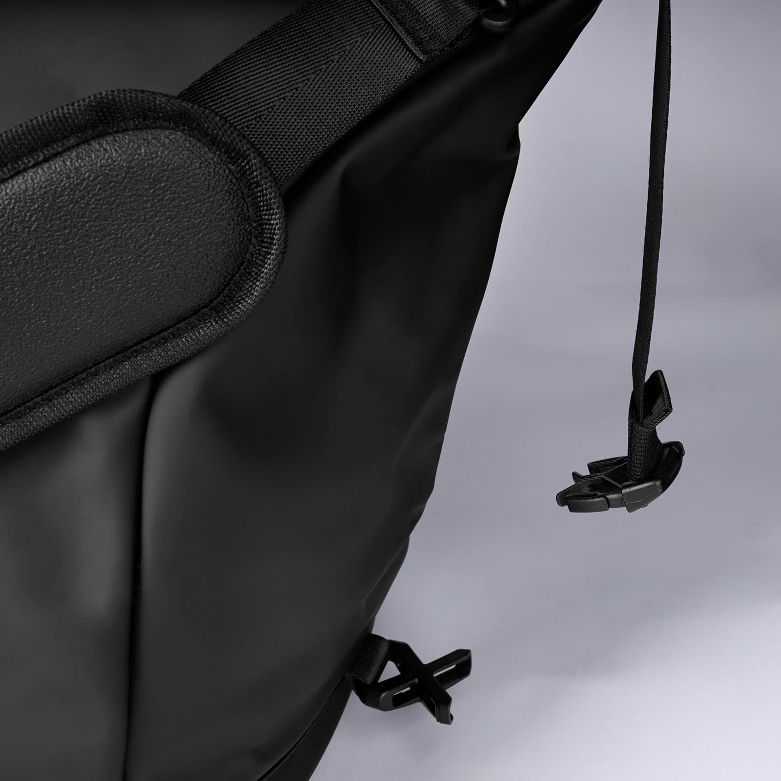 Nahaufnahme einer schwarzen Tasche mit verstellbarem Riemen, gepolstertem Schulterbereich und einer Kunststoffschnalle. Die Tasche scheint aus einem glatten, strapazierfähigen Material – möglicherweise Nylon oder Polyester – gefertigt zu sein und ist vor einem einfarbigen Hintergrund zu sehen.
