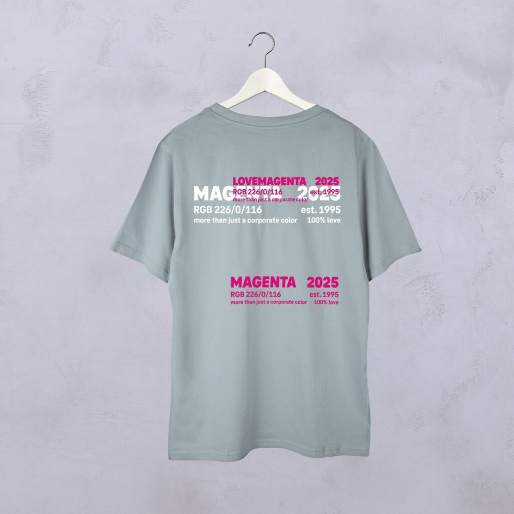 Auf einem hellgrauen T-Shirt, das an einem Kleiderbügel hängt, sind auf der Rückseite magentafarbene und weiße Schriftzüge zum Thema Magenta zu sehen, darunter „LOVEMAGENTA“, „2025“, „est. 1995“ sowie RGB-Farbcodes.