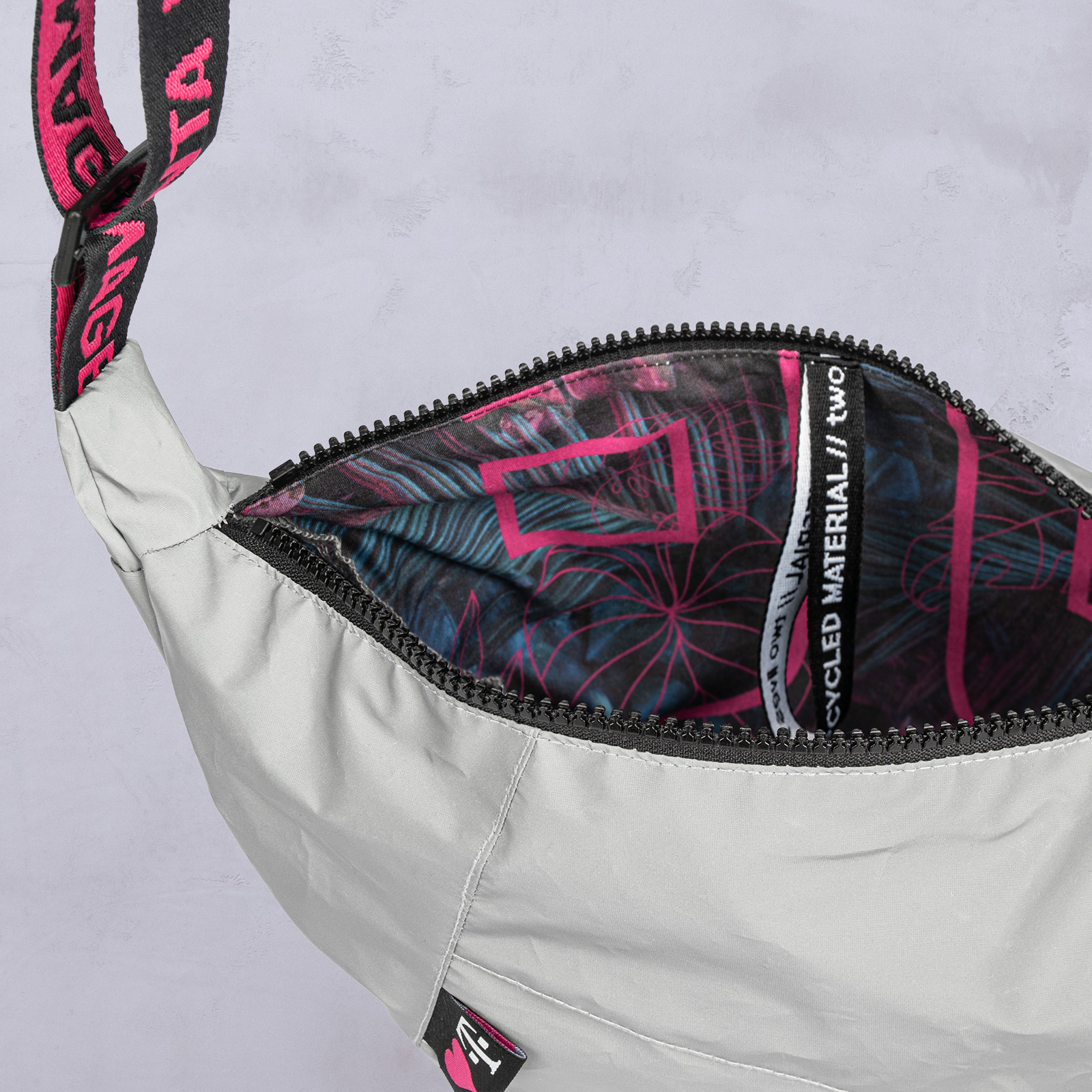 Eine graue Umhängetasche mit schwarzem Reißverschluss, deren Innenfutter ein buntes Muster in Rosa und Blau aufweist und deren Riemen mit rosa Schrift verziert ist.