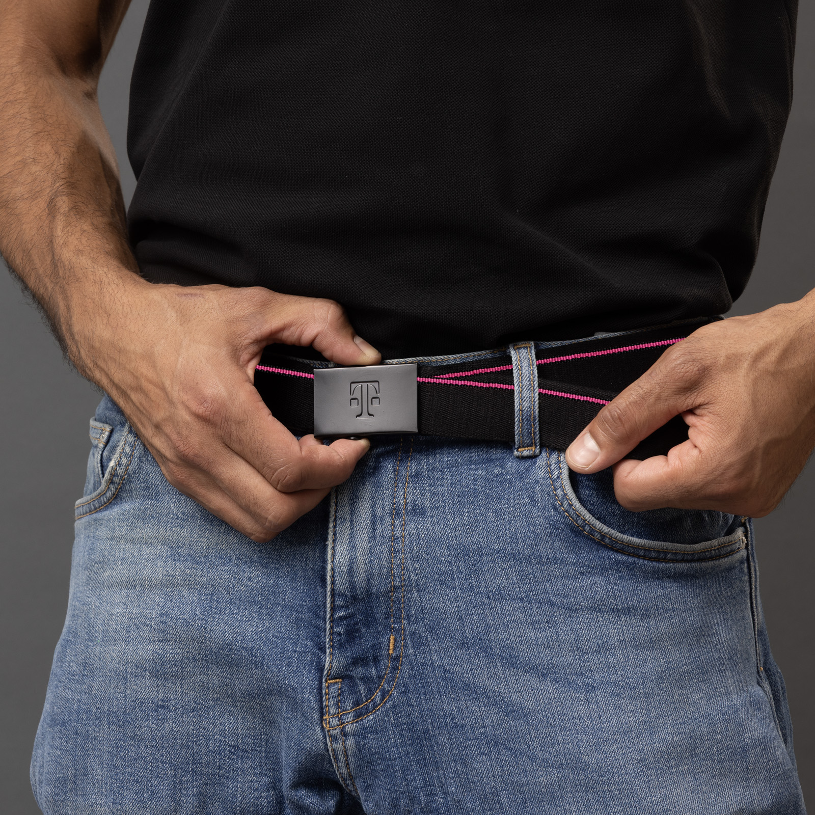 Eine Person in blauen Jeans und einem schwarzen T-Shirt richtet vor einem dunklen Hintergrund einen schwarzen Gürtel mit rosa Ziernähten und einer Metallschnalle mit einem T-Logo-Motiv zurecht.