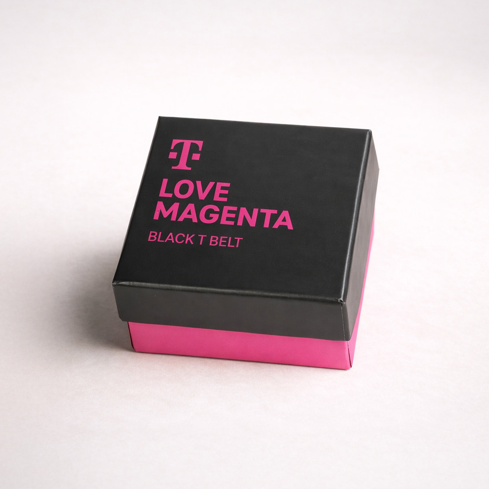 Eine schwarz-magentafarbene Geschenkbox mit dem T-Mobile-Logo und der Aufschrift „LOVE MAGENTA BLACK T BELT“ auf dem Deckel. Die Seiten der Box sind magentafarben, während der Deckel schwarz ist.