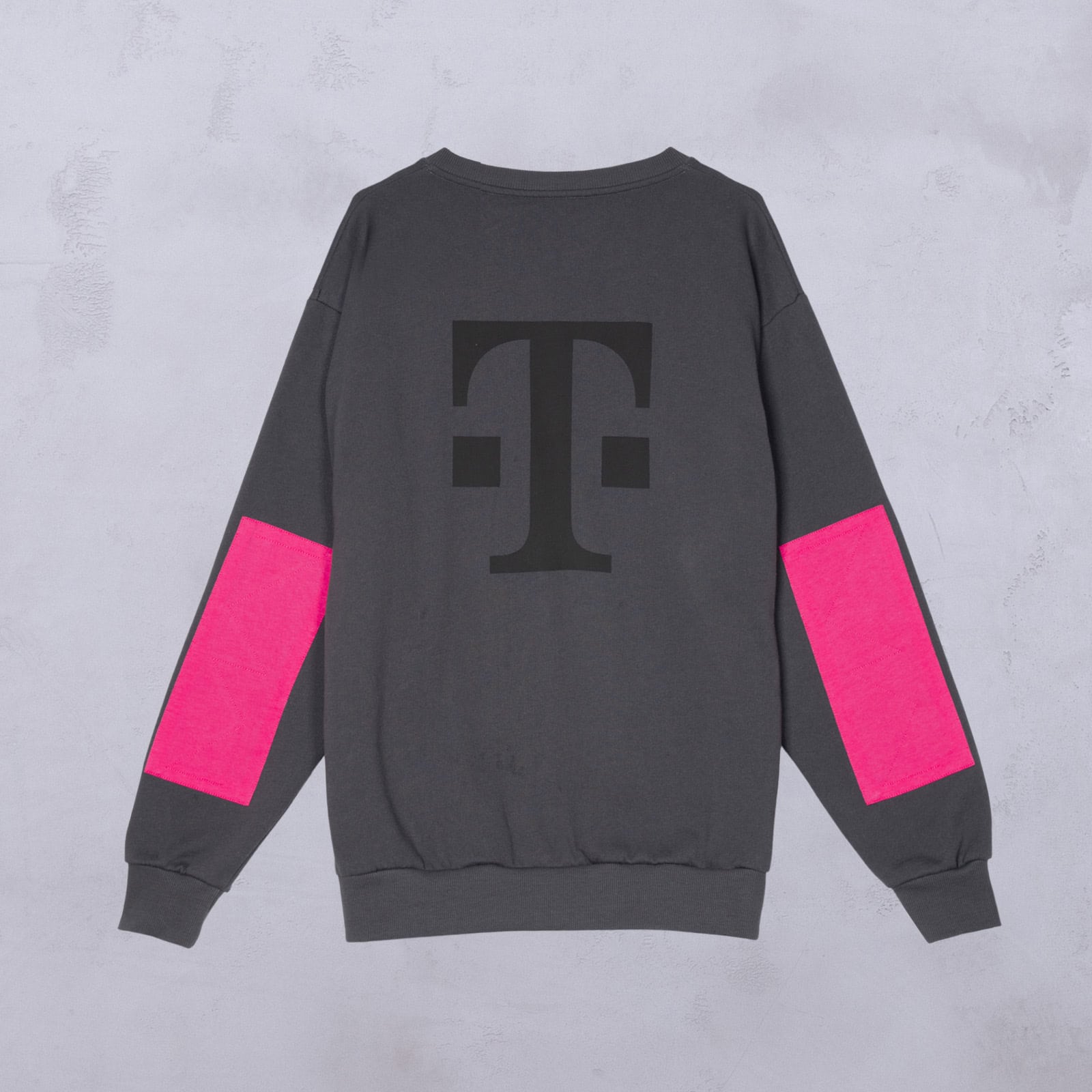Ein dunkelgraues Sweatshirt mit einem großen schwarzen T-Logo auf dem Rücken und leuchtend pinkfarbenen rechteckigen Aufnähern auf beiden Ärmeln, fotografiert vor einem hellgrauen, strukturierten Hintergrund.