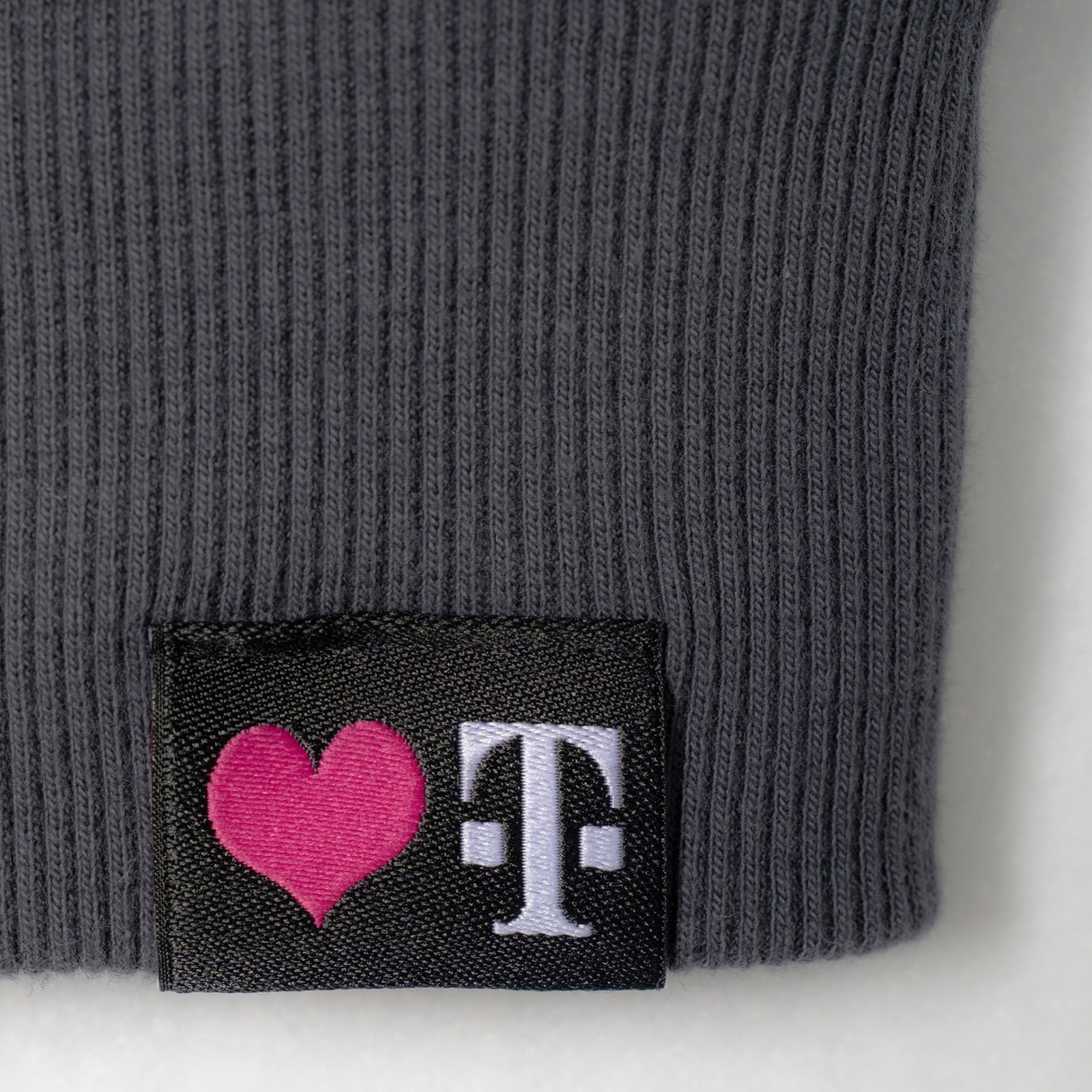 Eine Nahaufnahme eines grauen, gerippten Stoffes mit einem schwarzen Aufnäher, auf dem ein rosa Herzsymbol und ein weißer Buchstabe „T“ zu sehen sind.